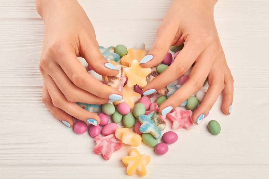 Female hands and colorful candies. Woman manicured hands and multicolored tasty candies on white wooden background close up.