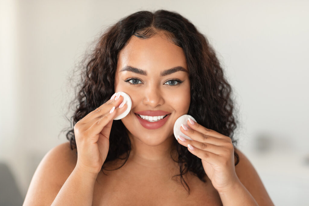 Happy black plus size lady holding cotton pads and smiling at camera, using cleansing lotion or hydrating toner, enjoying face care routine. Makeup remover. makeup remover types