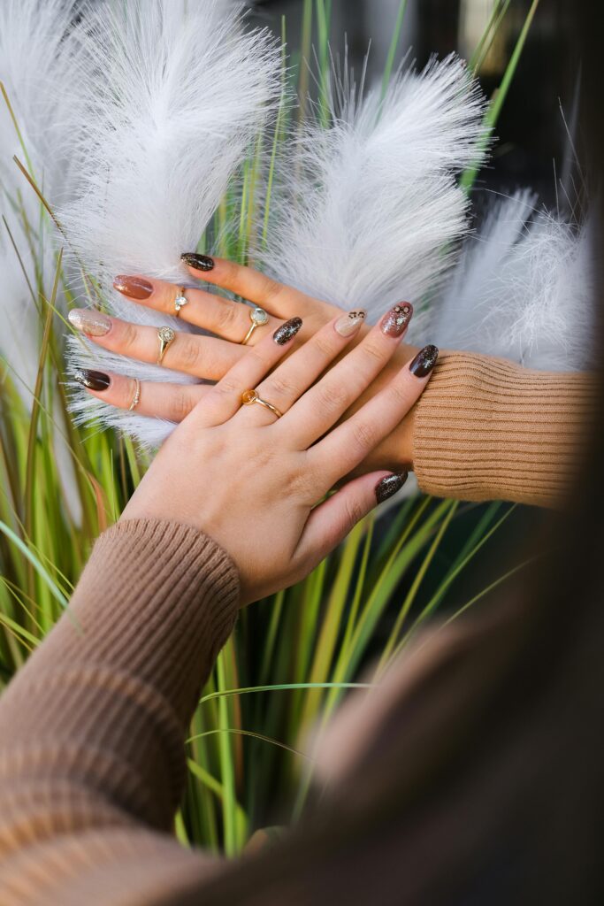 A woman with faceless features showcasing nails painted in various shades of brown with glitter accents and intricate designs.