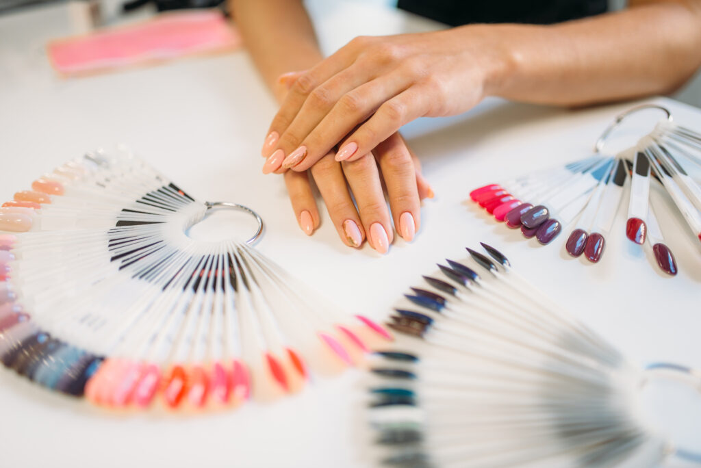 Well-groomed female hands against nail sample fan on white table. Manicure art designs set, colorful collection in beauty salon. Home nail designs. DIT nail designs.