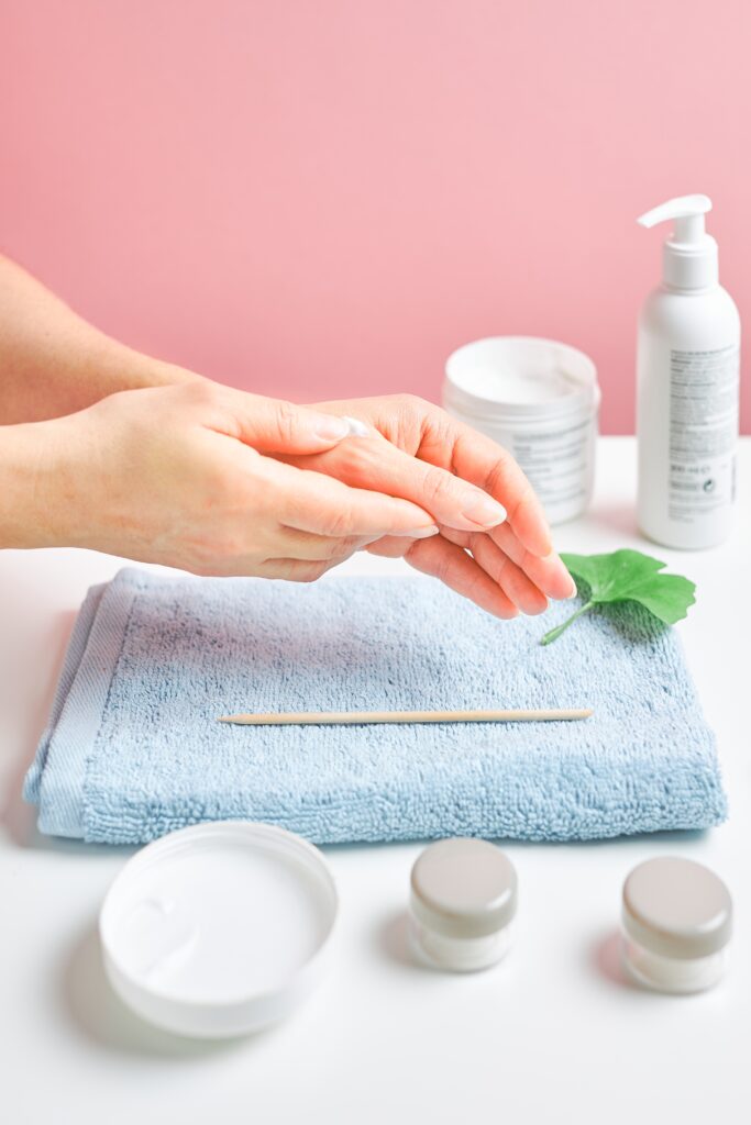 Woman applying  cosmetic moisturizing hand cream. Cosmetic products, green leaves on white table. Spa, manicure, skin care concept. Flat lay, overhead view. nail care. nail treatments. Home care.