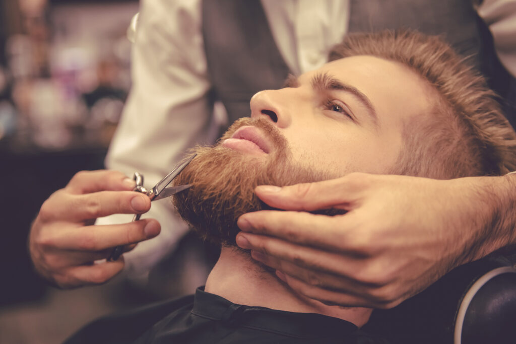 Side view of handsome bearded man having his beard cut by hairdresser at the barbershop.