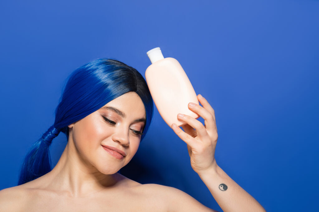 beauty trends, body and hair care concept, portrait of tattooed young woman with vibrant hair color posing with bare shoulders on blue background, holding cosmetic bottle with shampoo, advertisement. hair masks for color-treated hair