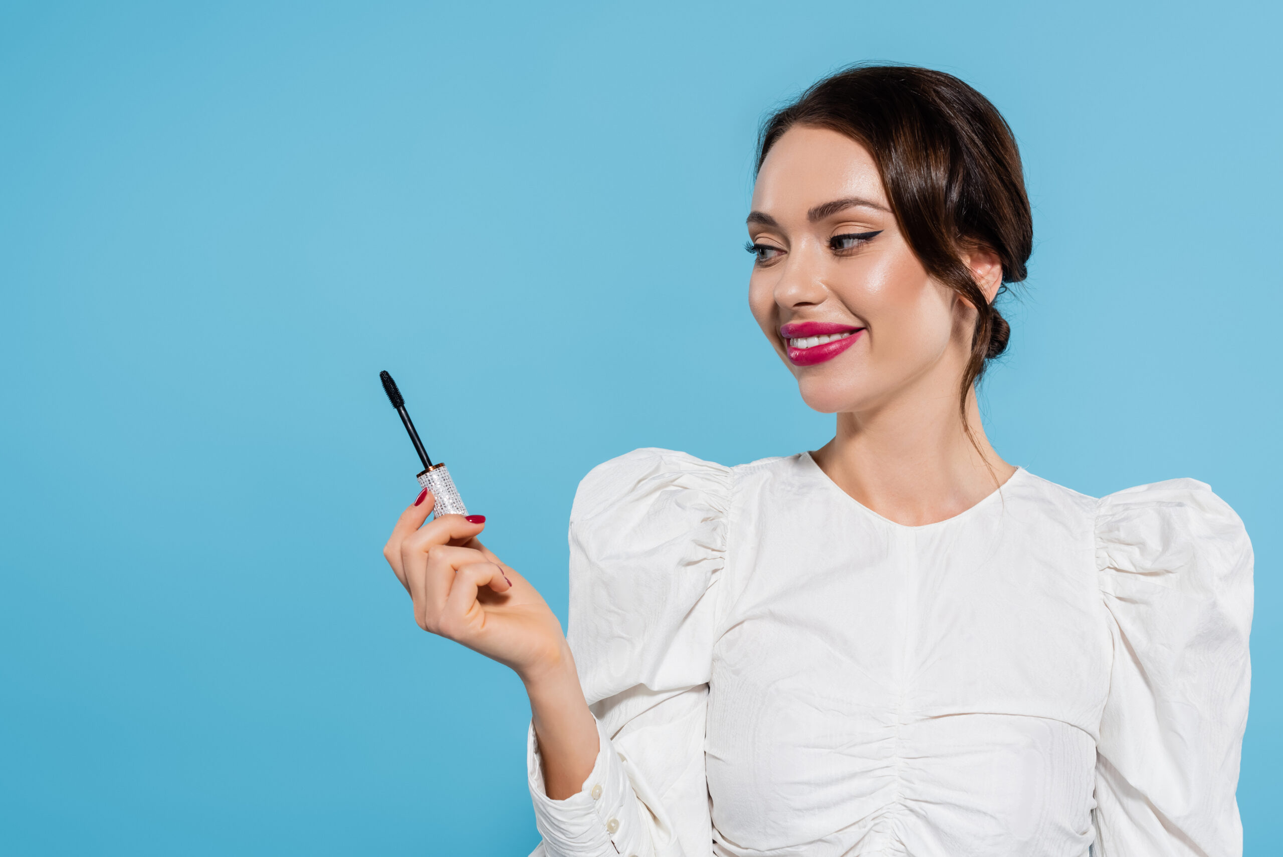 cheerful young woman in white blouse holding mascara brush isolated on blue