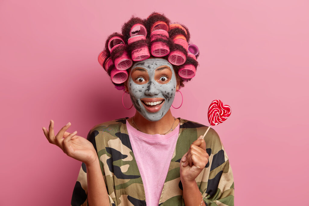 Fabulous woman looks with happy expression, raises hand and holds delicious tasty lollipop wears hair rollers, makes perfect hairstyle smiles broadly dressed in khaki dressing gown beauty mask on face. care. treatment. DIY