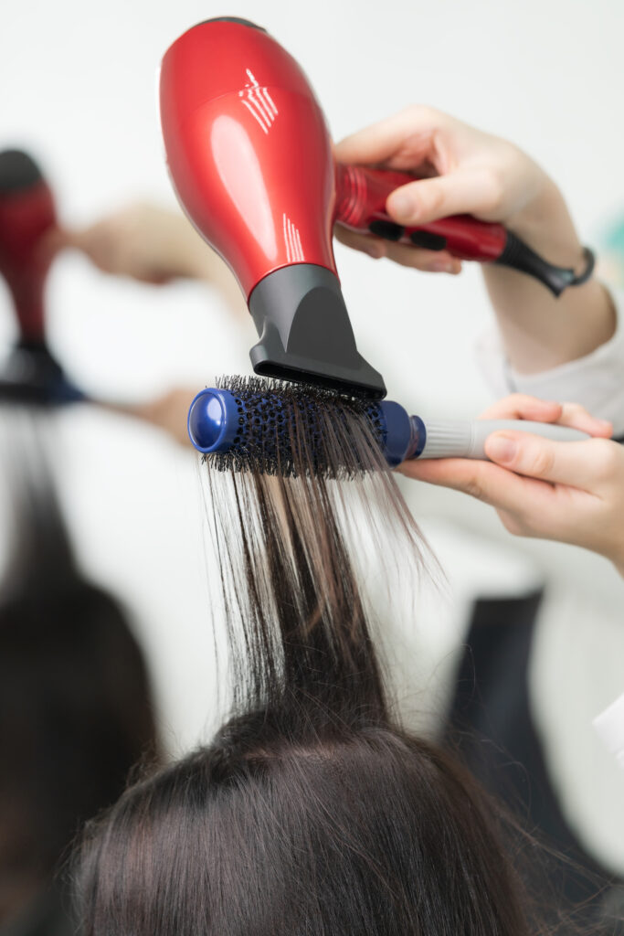 Hands of hairstylist dries long brunette hair of client using red hairdryer and blue comb.