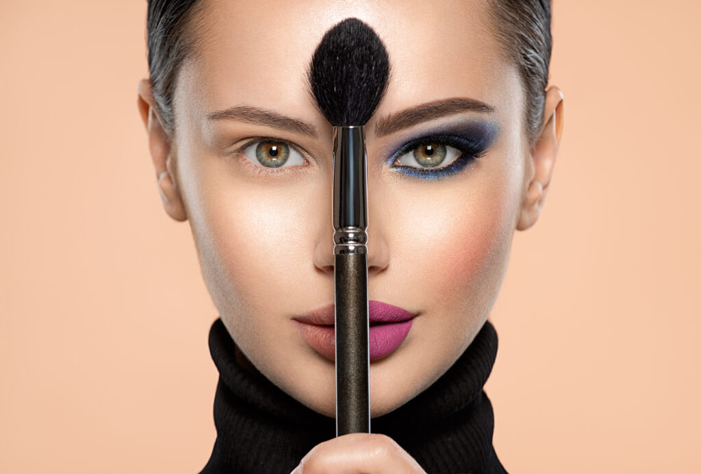 Portrait of a girl with cosmetic brush at face. Woman holds a big makeup brush right in the middle of the face. One half face of a beautiful white woman with  bright makeup and the other is natural.