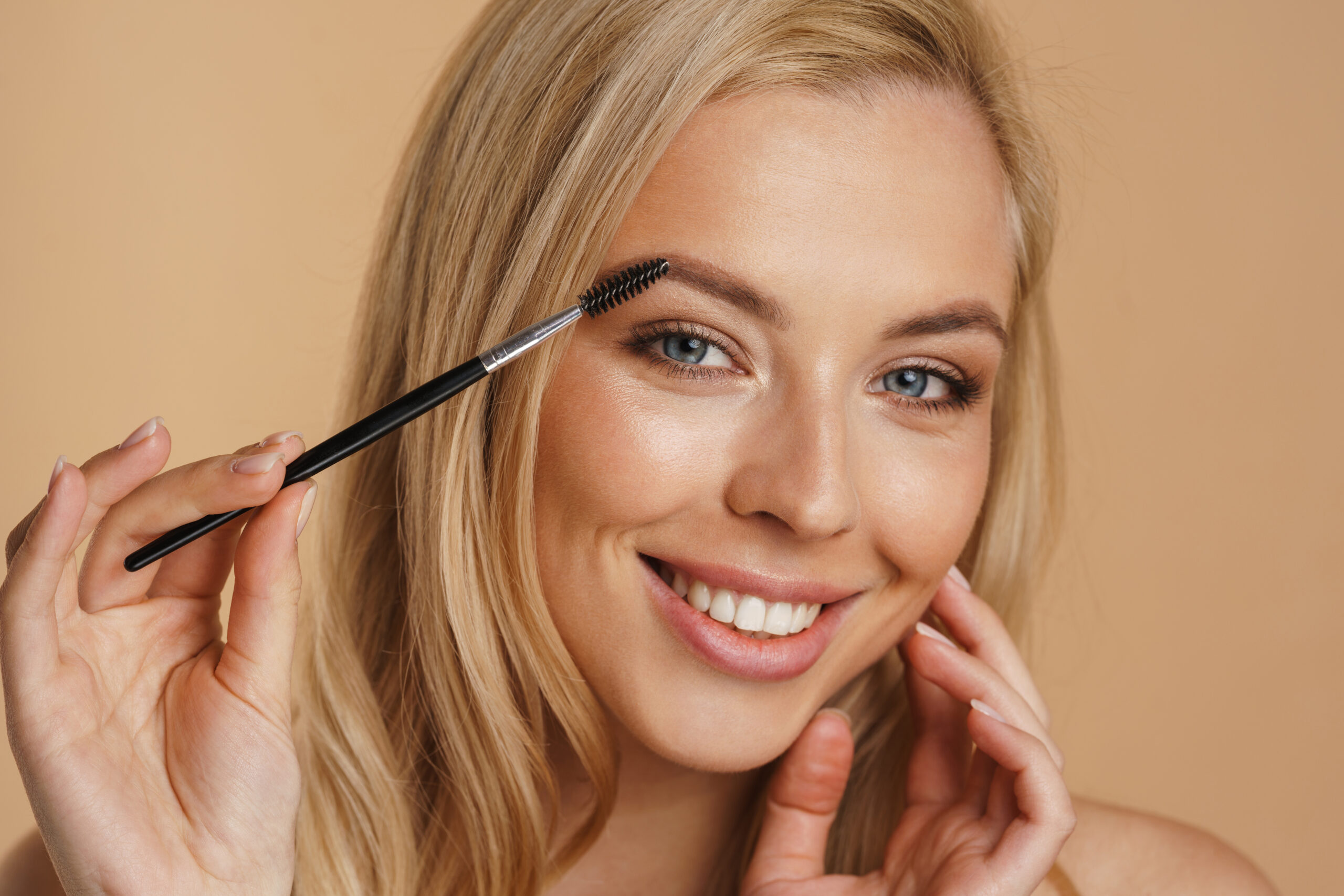 Smiling young white blonde woman with bare shoulders standing over beige background, holding eyebrow brush. eyebrow coloring.