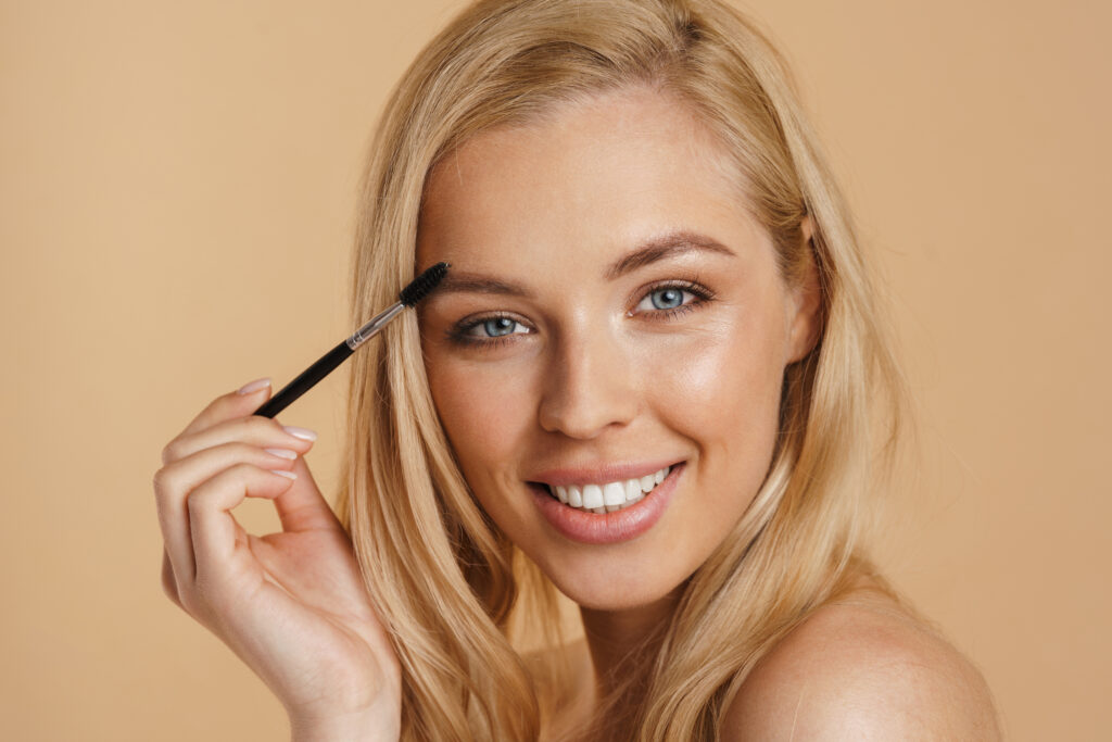 Smiling young white blonde woman with bare shoulders standing over beige background, holding eyebrow brush. eyebrow coloring methods