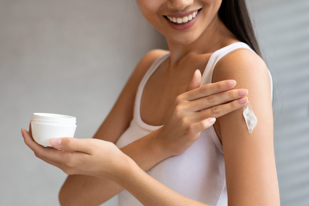 Unrecognizable brunette woman in white top applying skin butter or mosturizer on her shoulder after morning shower or bath and smiling, holding jar with cream, cropped, copy space. body cream. skin care