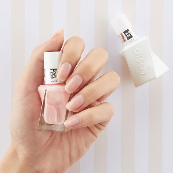 A faceless woman holding the Essie Gel Couture gel polish bottle in a light pink shade, with a white gel polish bottle beside her hand. Her nails are painted in the same light pink shade.