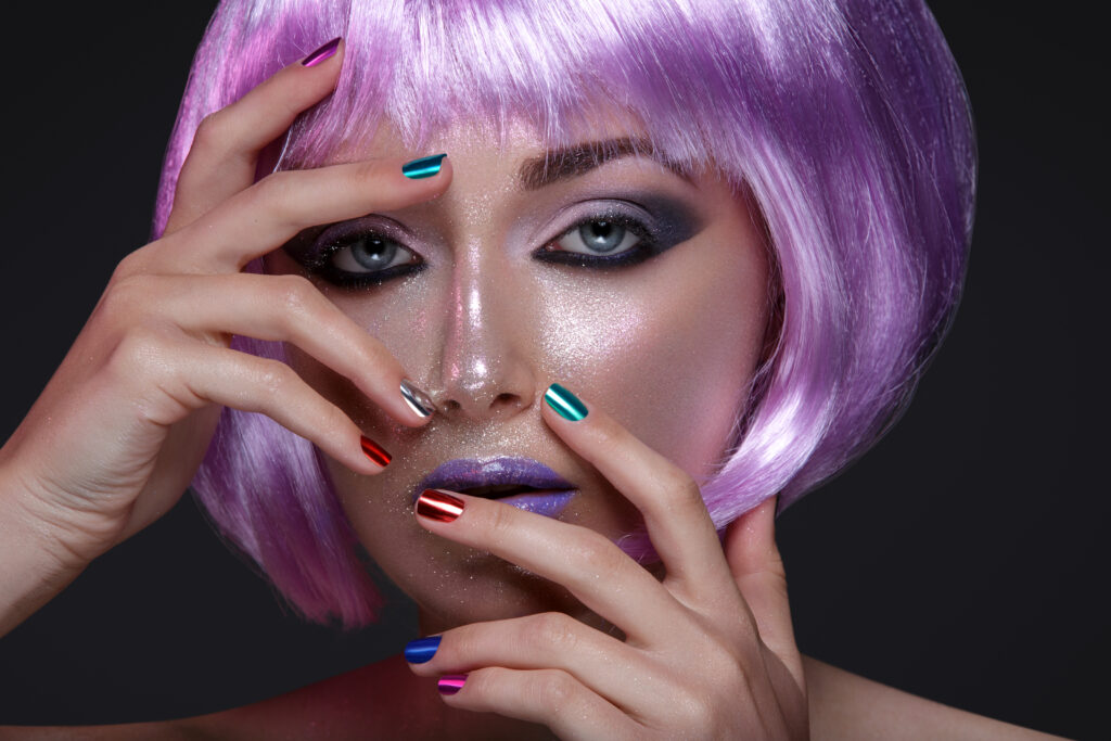 Beautiful young woman in purple wig with artistic violet lipstick and eye makeup with pink sparkles. Beauty shot on black background. Colorful nails. nail effects. woman earing colorful nail effects