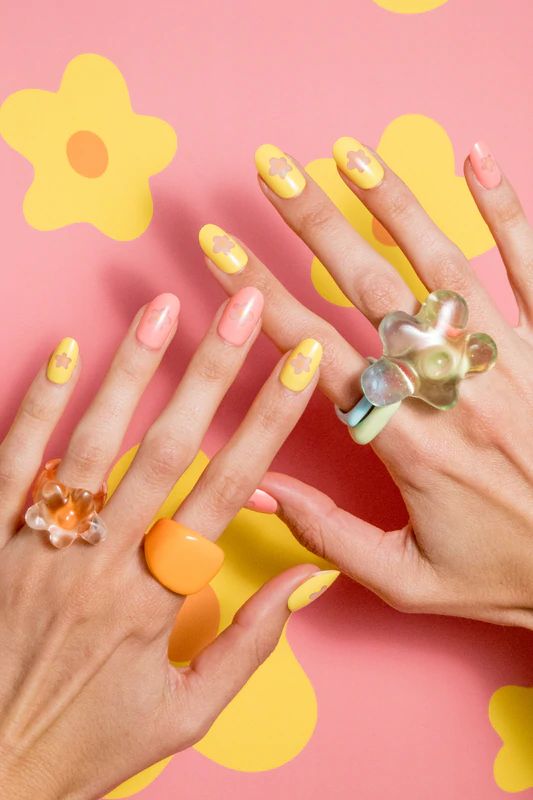 A faceless woman with nails painted in pink and yellow, featuring clear star design, wearing vibrant rings on a yellow and pink background.