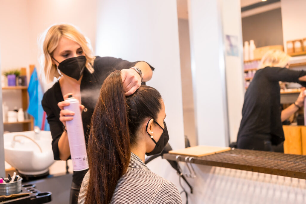 A hairdresser in a protective mask spraying client's hair with hairspray