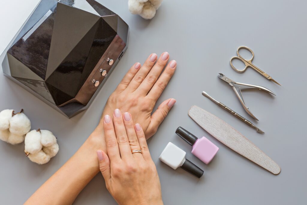 A faceless woman is preparing tools for applying Harmony Gelish gel polish. The setup includes a nail file, an LED nail lamp, two bottles of gel polish in different shades, nail scissors, and various other nail tools.