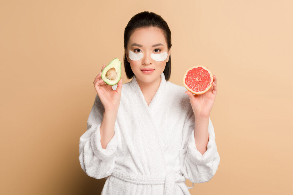 beautiful asian woman in bathrobe with eye patches on face holding grapefruit and avocado halves on. eye care. eye caring. take care for the skin around the eye. eye masks.