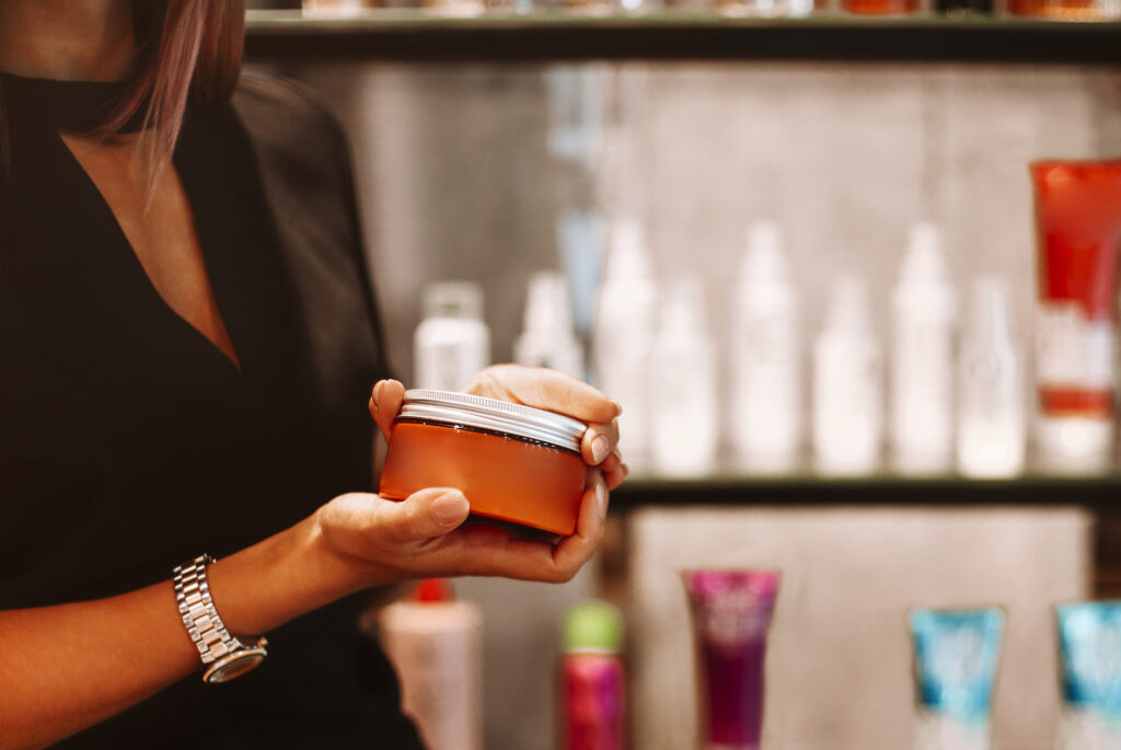 Cropped photo of female hairdresser holding jar of hair mask, woman coiffeur showing special styling product stands against blurred background of modern beauty salon. Haircare and cosmetic concept