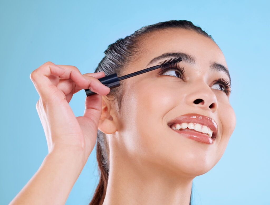 Studio shot of an attractive young woman applying mascara against a blue background. Lash serum. Long lashes.