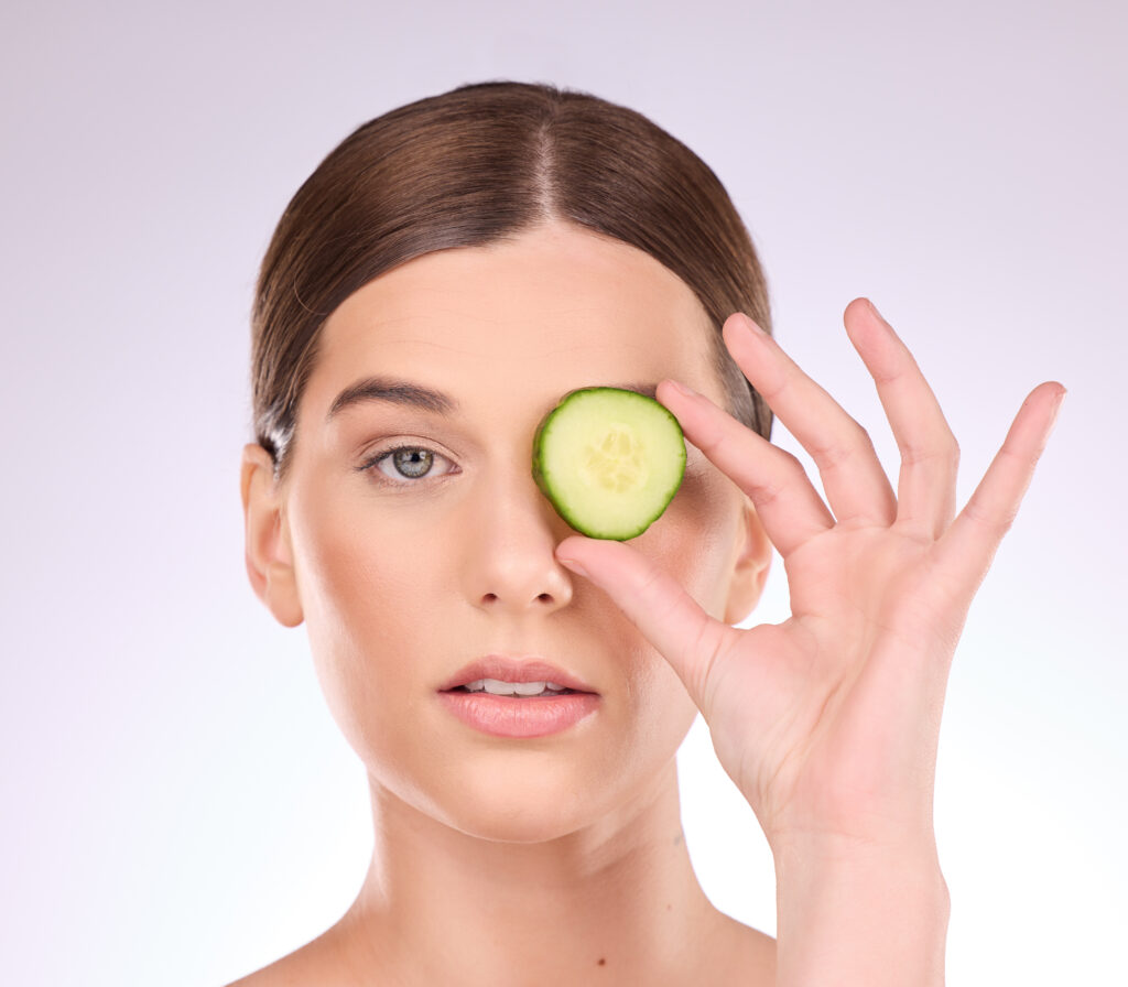 Woman, face and hand with cucumber for skincare nutrition or healthy diet against a gray studio bac