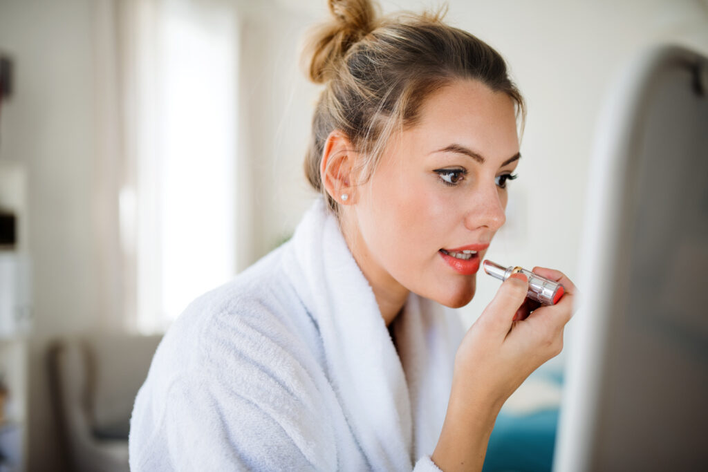 A young woman indoors at home in the morning, putting on lipstick. Alternatives to Lip Botox.