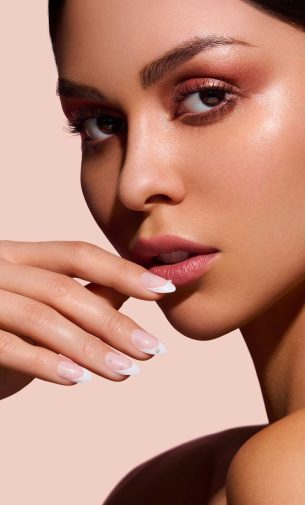 A beautiful woman looking to the side, wearing nude makeup and showcasing classic French nails adorned with rhinestones. French nail trends emphasize the timeless elegance of this chic style.