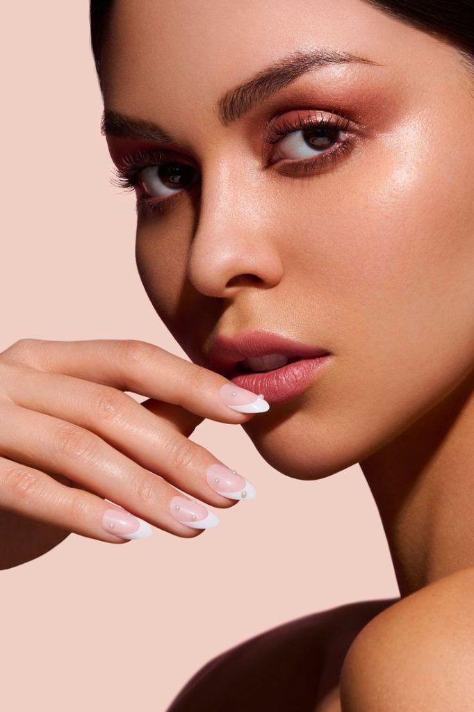 A beautiful woman looking to the side, wearing nude makeup and showcasing classic French nails adorned with rhinestones. French nail trends emphasize the timeless elegance of this chic style.