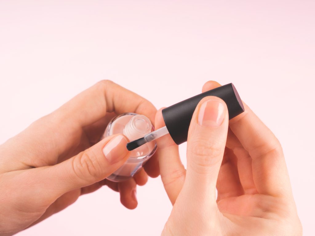 Applying nude nail polish to woman's hand over delicate pink background. Close up. Top Coats