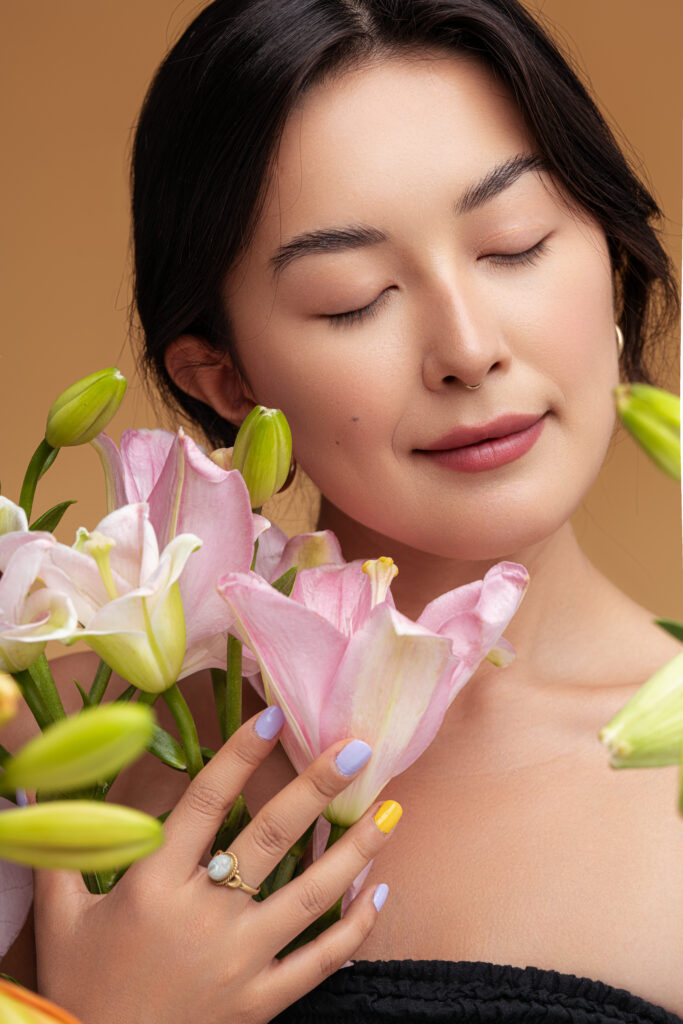 Young Asian female with closed eyes touch and enjoying smell of natural lily flowers against brown background. Nail Trends for 2025