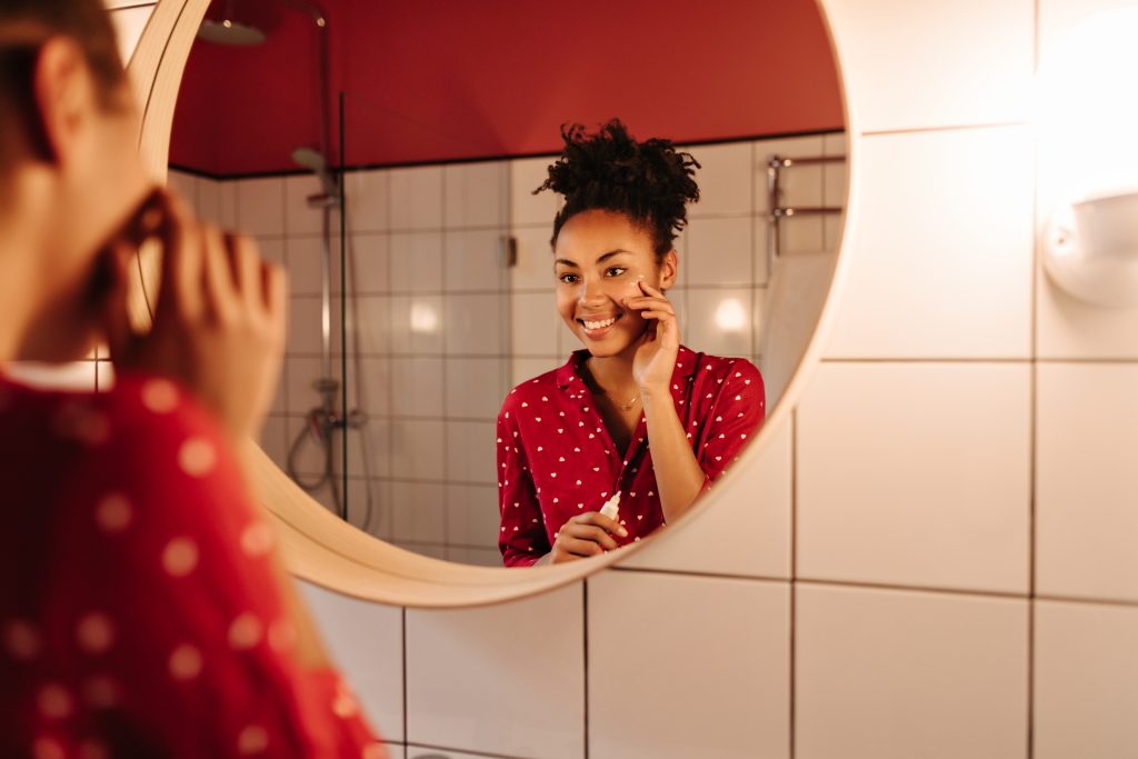 all-day care: from hair to toes! Brunette girl in red shirt does morning routine in bathroom and moisturizes her face with cream. home. DIY. caring.