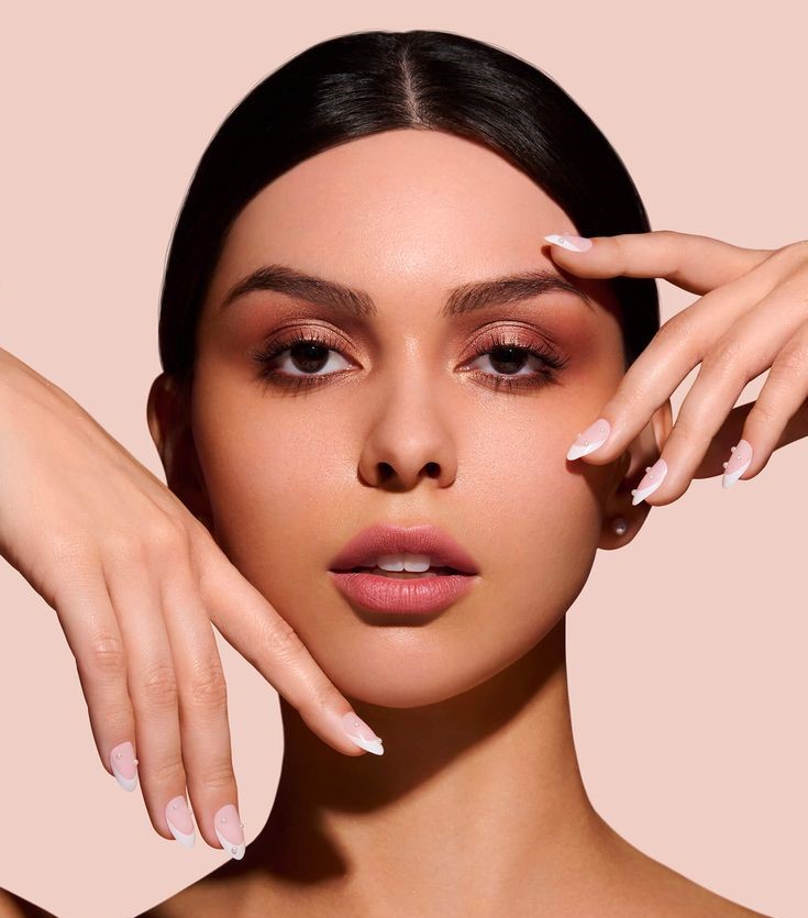 A beautiful woman wearing nude makeup, showcasing classic French nails adorned with rhinestones. French nail trends highlight the timeless elegance of this stylish look.