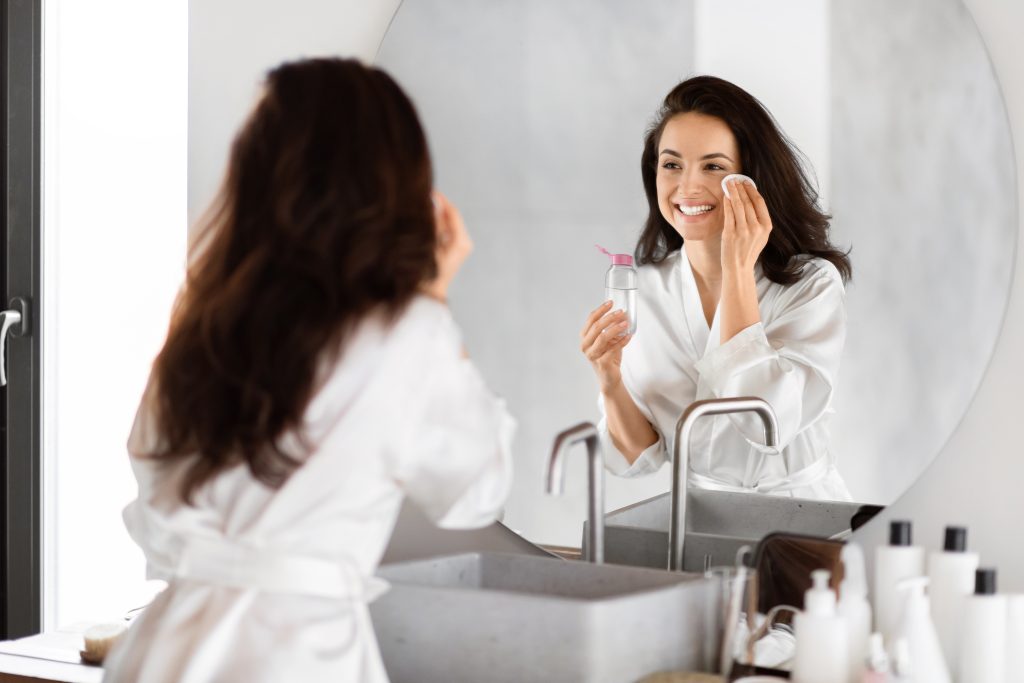  makeup removers. makeup remover types. liquid makeup remover. In a bathroom, a joyful millennial woman uses a cotton pad on her face as a part of her skin care routine