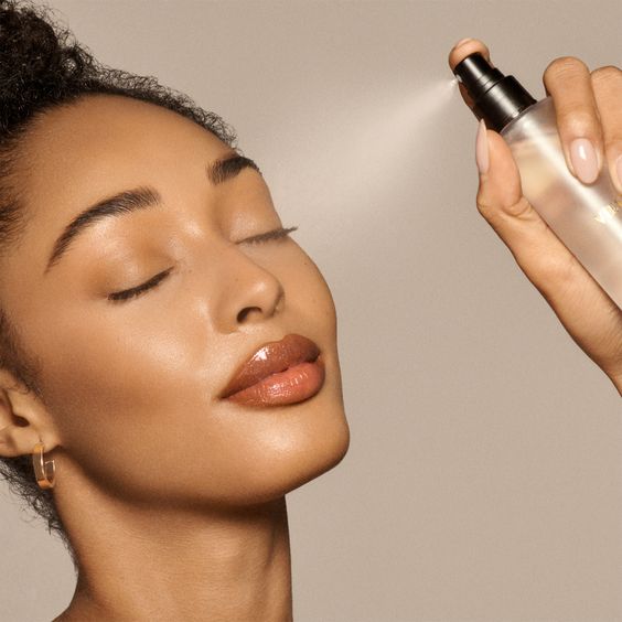 A gorgeous woman with flawless, professional makeup is spraying setting spray on her face in front of a gray background.