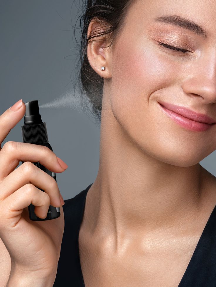 A smiling woman with minimal makeup is applying setting spray to her face against a blue background, showcasing her light orange manicure.