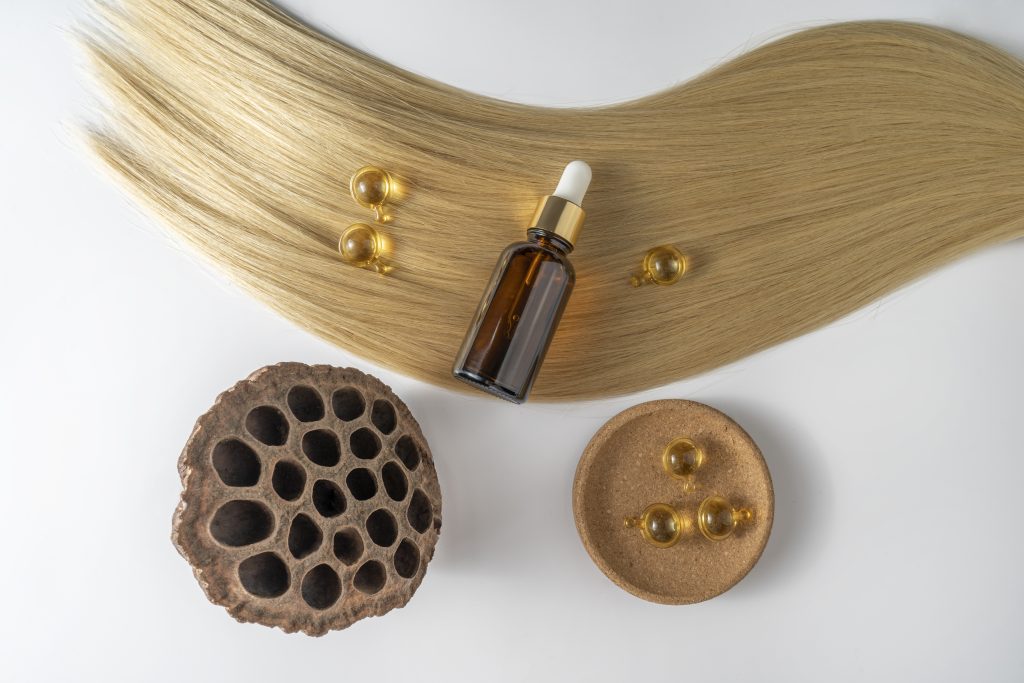 Growing Out Short Hair at Home. Nourish Your Hair from the Inside Out. A hair care oil or serum in a brown dropper bottle and golden capsules lying on a blond hair strand, product marketing mockup.