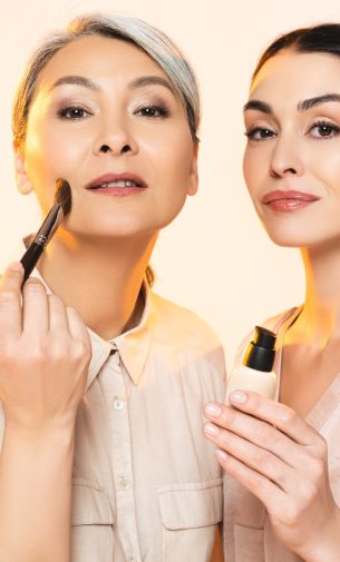 beautiful multicultural women holding cosmetic brush and makeup foundation isolated on beige