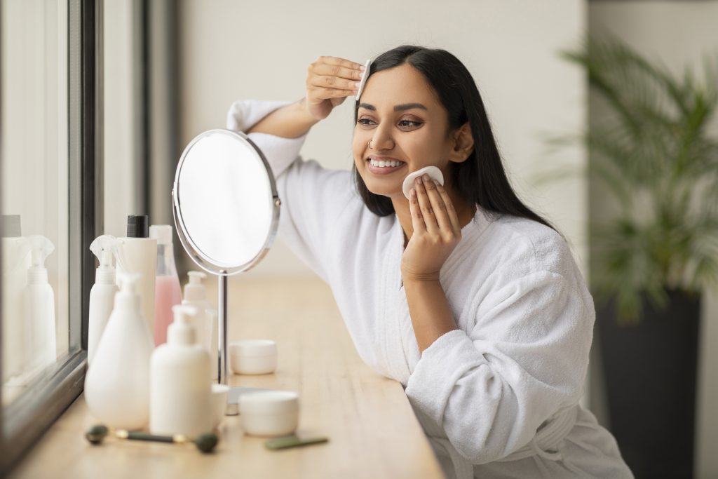 How to Remove Foundation. Beautiful Smiling Indian Woman Cleansing Skin With Cotton Pads While Making Daily Beauty Routine In Bathroom, Looking At Mirror, Attractive Young Lady Enjoying Home Skincare Treatments, Copy Space. remove makeup. makeup remover.