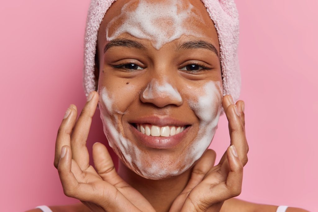 Beauty and cosmetology concept. Studio shot of young pretty smiling happy European lady applying soap on face as part of daily routine procedures holding hands near cheeks posing on pink background