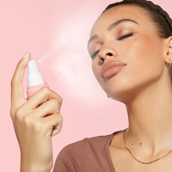 A beautiful woman with stunning, professional makeup is applying setting spray to her face against a pink background.