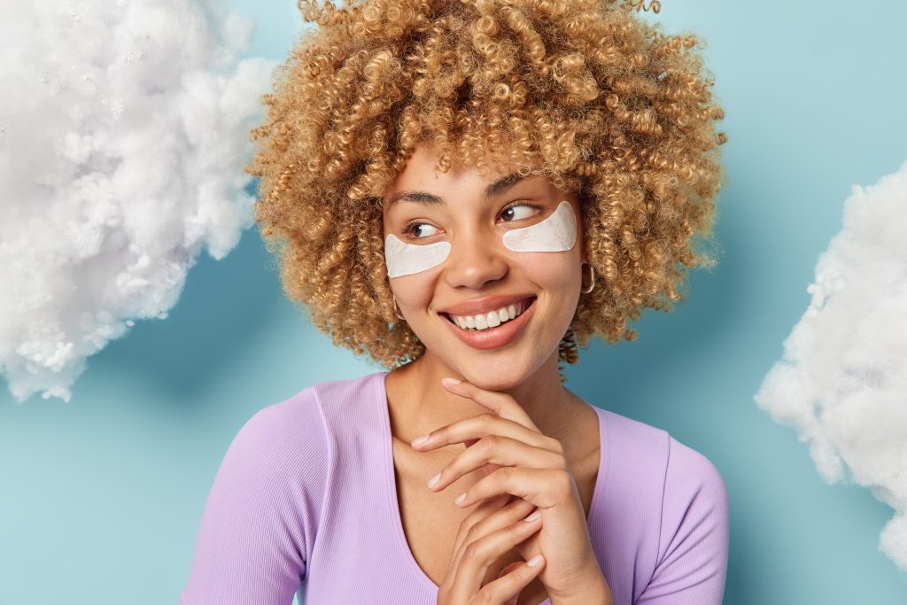 eye masks. eye mask. eye care. home eye caring. Charming curly haired young woman touches chin gently smiles toothily looks aside applies beauty patches under eyes dressed in purple jumper isolated over blue background white clouds overhead.