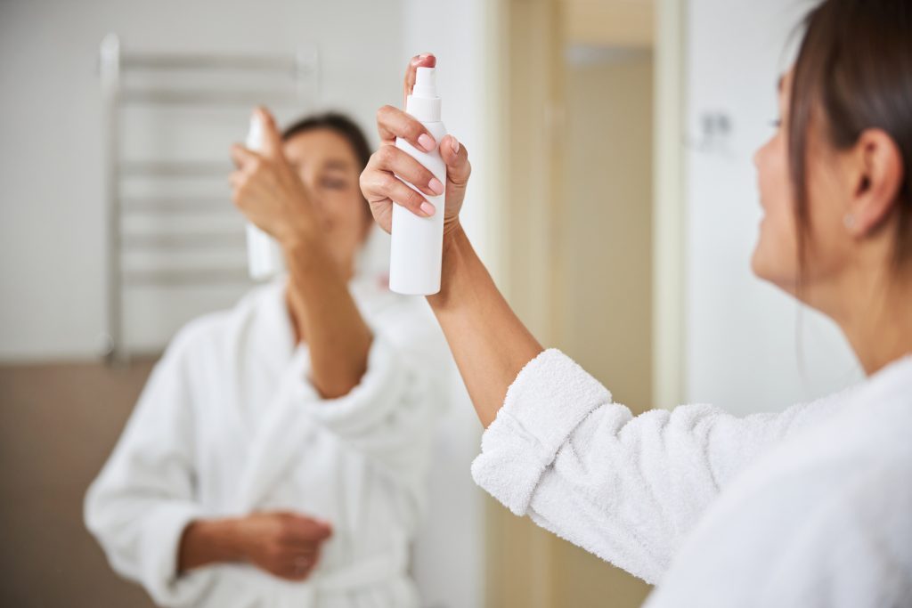 Cropped head back view portrait of beautiful elegant woman hand with white bottle of skin spray in front of the mirror in bathroom. makeup setting spray. setting spray. long-lasting makeup. makeup setting sprays.
