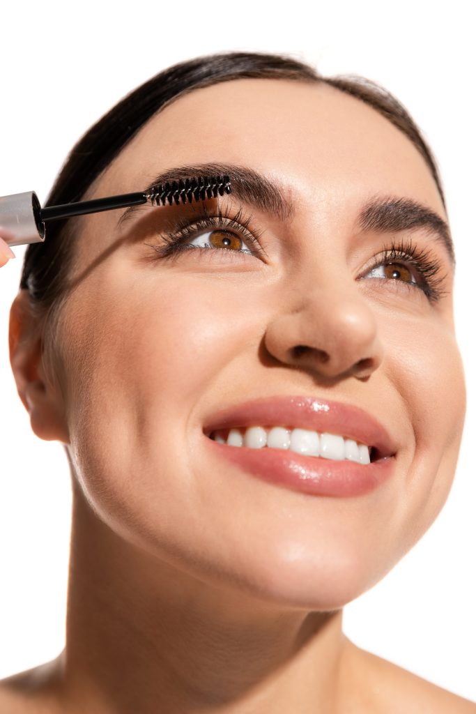 close up of cheerful woman applying mascara while looking up isolated on white