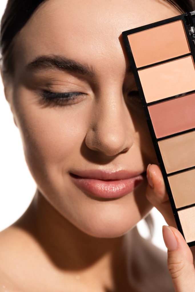 close up of smiling young woman covering eye with eye shadow palette isolated on white