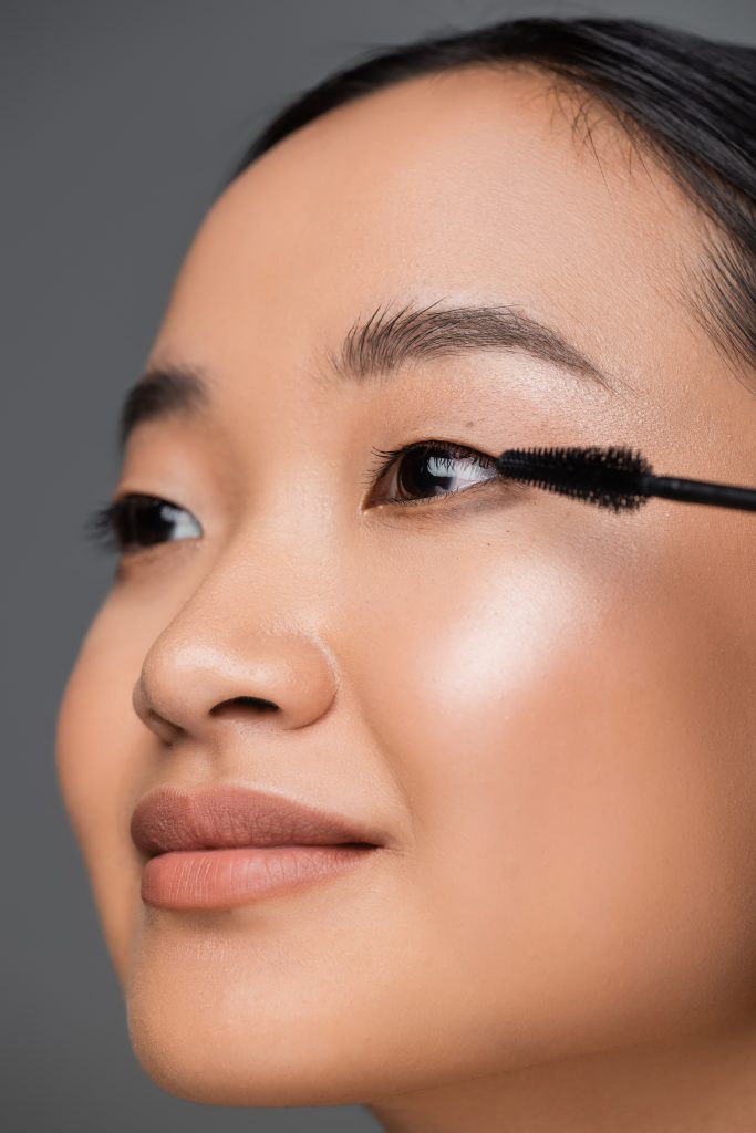 close up view of asian woman with makeup foundation applying black mascara isolated on grey