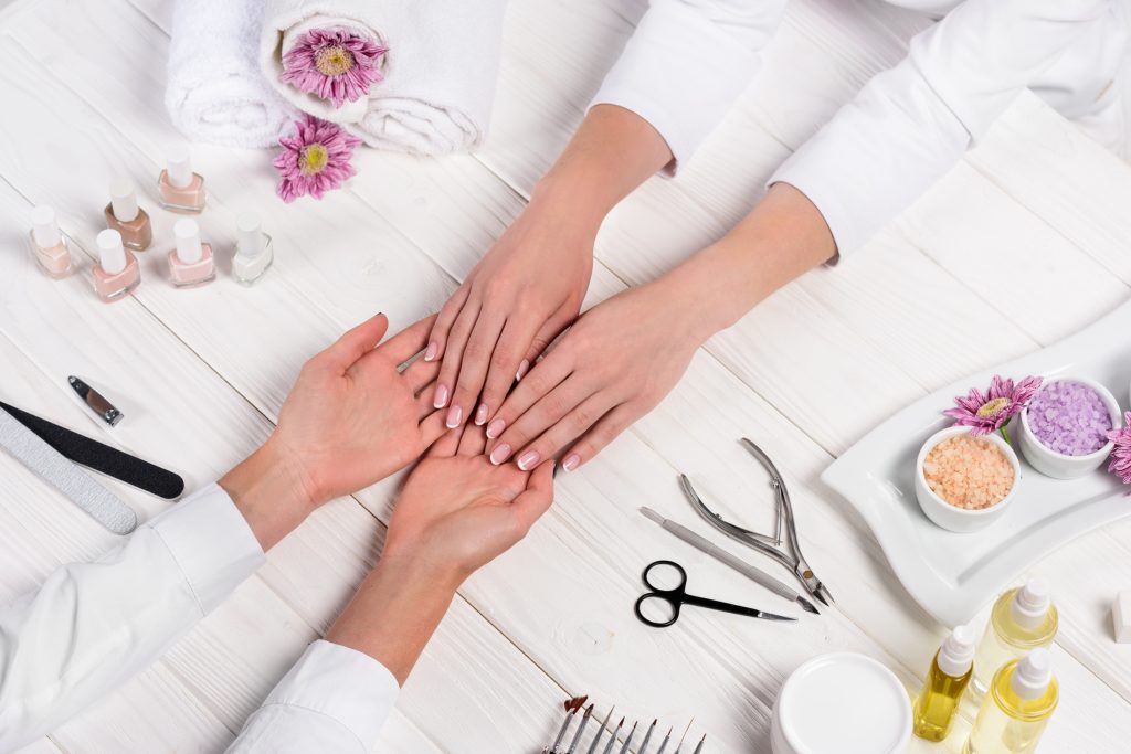 cropped image of manicurist looking at hands of woman at table with flowers, towels, nail polishes, nail care. home nail care. healthy nails.