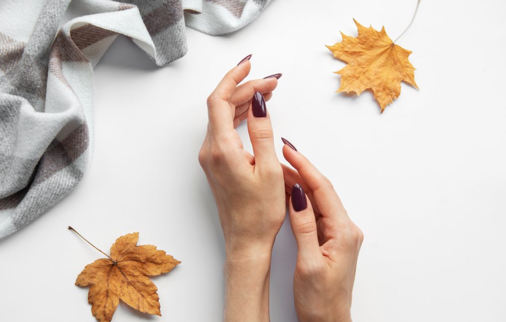 2024 Fall Nail Trends. A pair of hands with dark nail polish gently touch each other, surrounded by fallen autumn leaves and a soft, patterned blanket, evoking a cozy atmosphere.