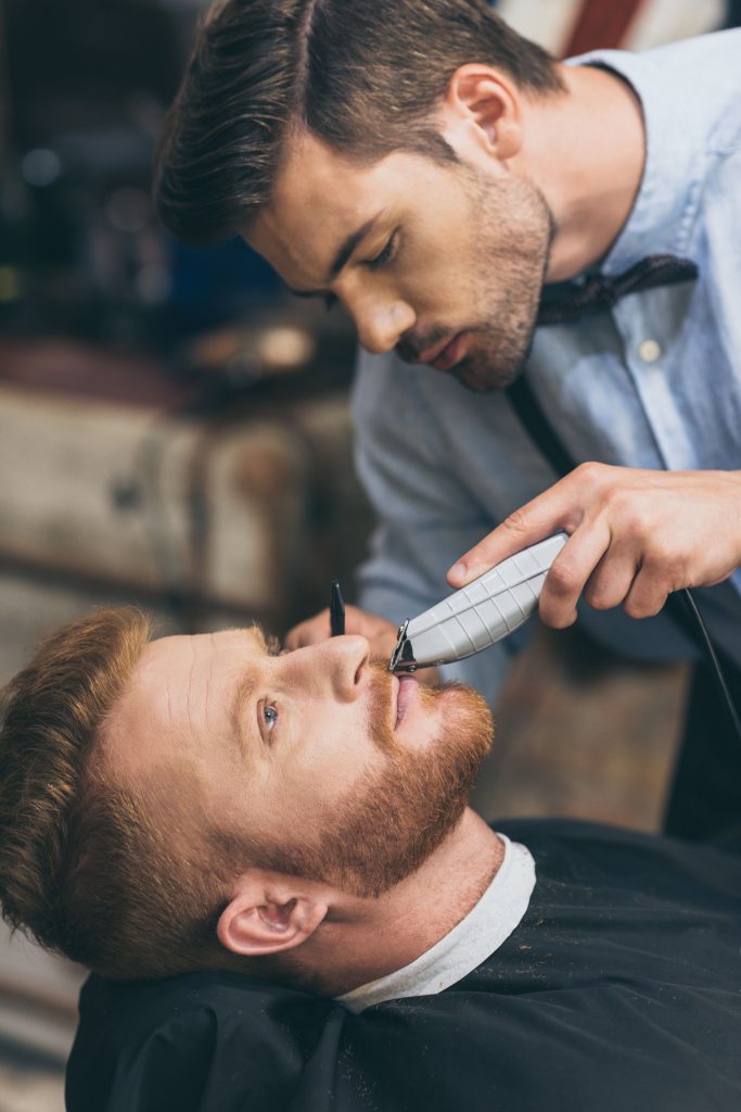 Barber trimming bearded man with shaving machine in barbershop. Hairstyling process. Close-up of a barber shaving the beard of a bearded male. Male barber trimming customers beard in barber shop.