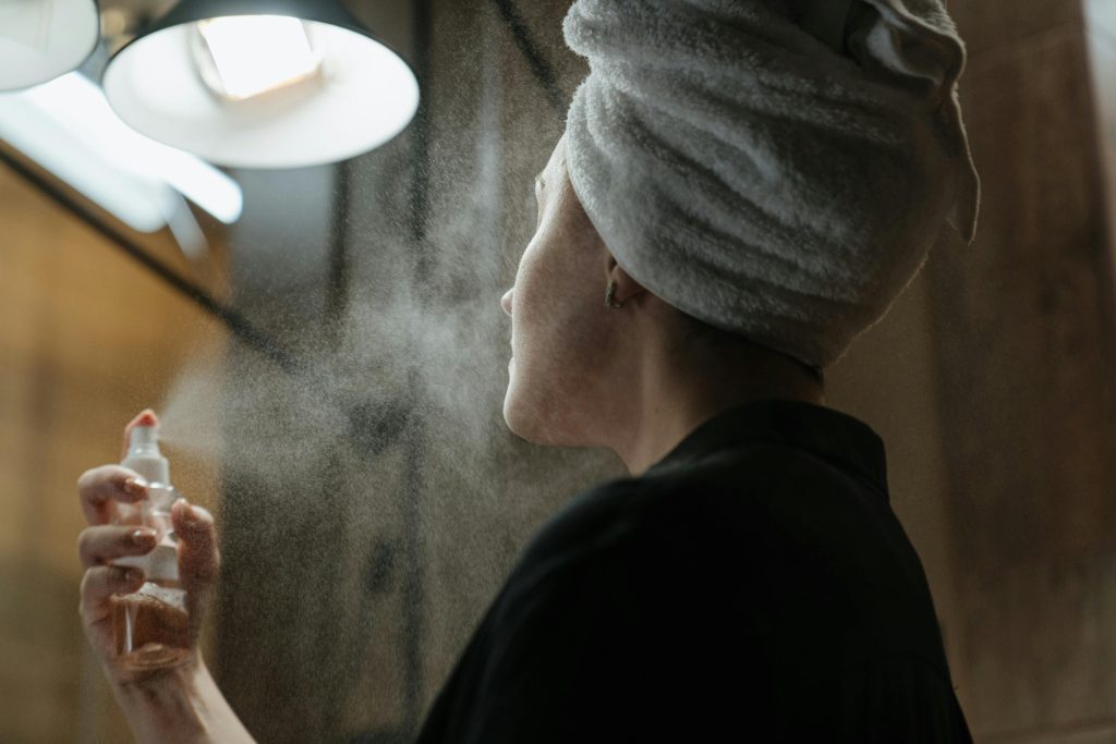 A faceless woman wearing a towel on her head is applying spray to her face.