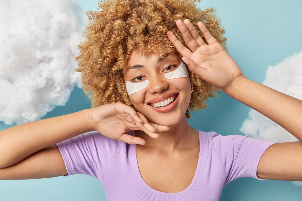 eye masks. Photo of curly haired woman touches face gently smiles toothily looks directly at camera dressed in casual purple t shirt applies white beauty patches under eyes poses against blue background. eye mask