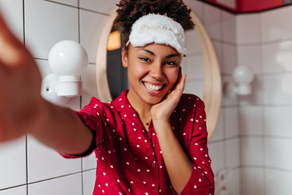 Charming dark-skinned woman in sleep mask is smiling, looking at camera and taking selfie. Common Night Skincare Mistakes to Avoid.