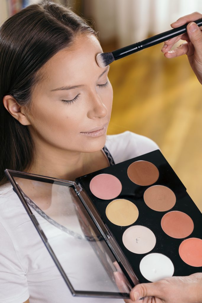 Make up artist working in a make up studio, applying the highlighters to female client's face. 